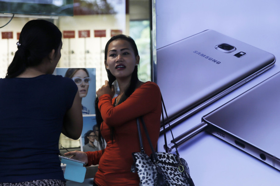 A woman holds her smartphone as she talks to a colleague near a Samsung Galaxy Note 5 advertisement at a mall in Jakarta